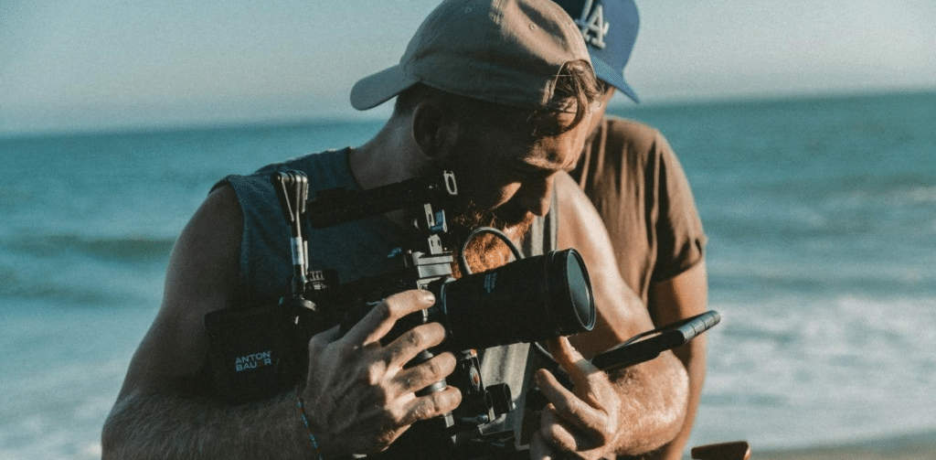 a man holding a film camera on a beach - he's peering into the viewfinder.
